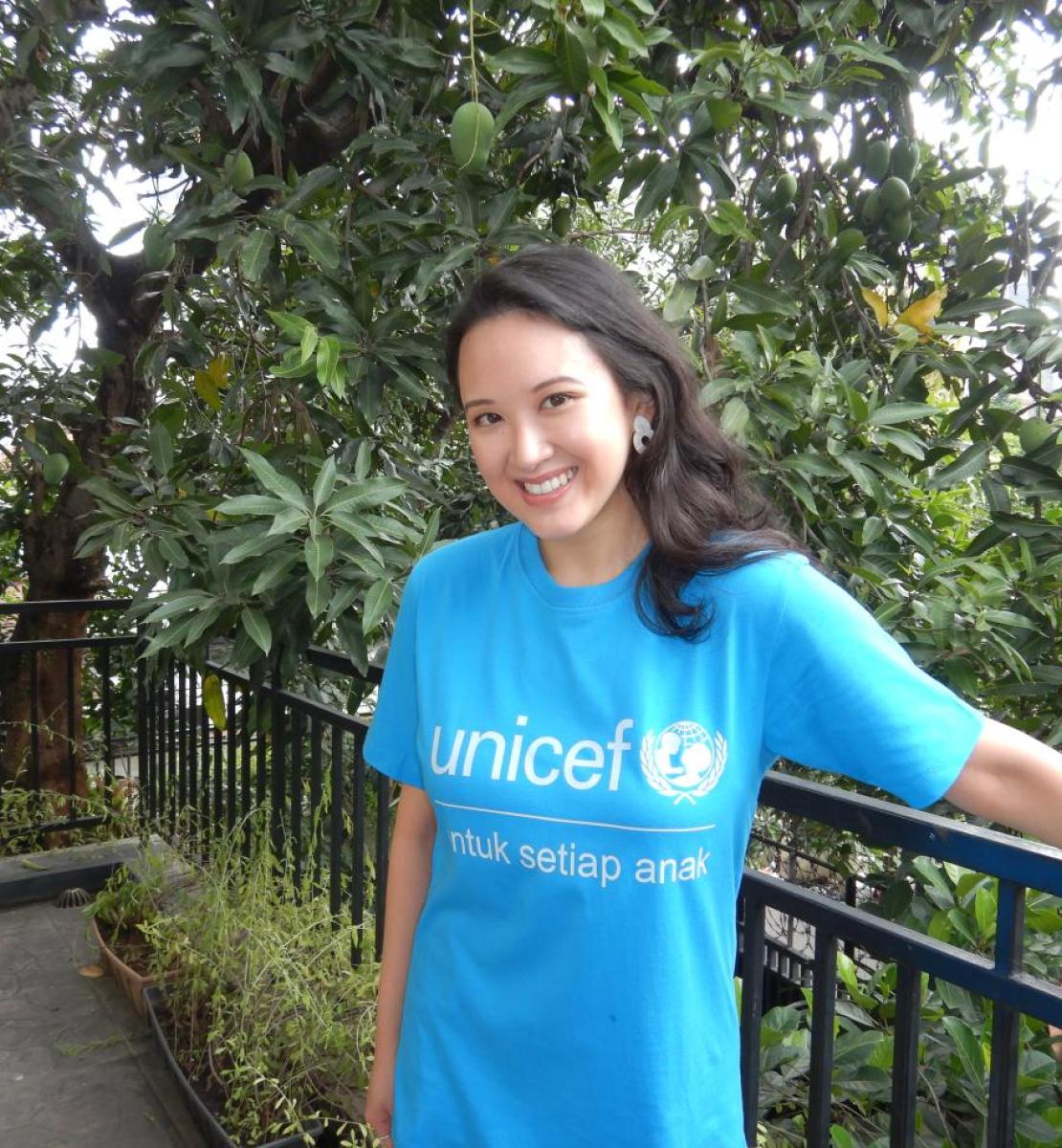 Una mujer mira con seguridad y sonríe a la cámara, usa una franela azul con el logo de UNICEF, y se encuentra en una espacio exterior, lleno de vegetación.