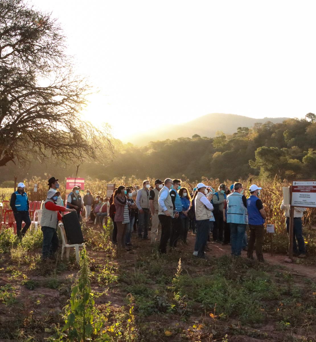 Un grupo de personas con mascarillas y hace fila en un área verde para visitar el centro de formación de demostración en la comunidad de Arenal, en Bolivia.