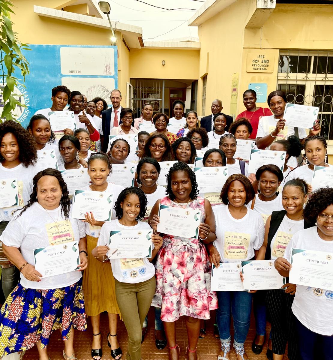 Women in white t-shirts hold certificates in their hands, in front of a municipal building, with the RC standing in the center-back.