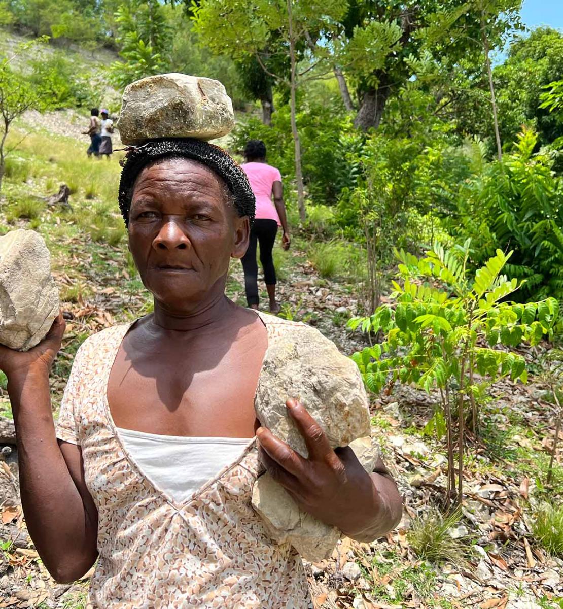 Portrait of a woman in a beige and white blouse, looking determinedly at the camera and she is part of a line of women carrying rocks in their hands and on their heads as they descend a ravine on a hillside in the southern Haiti.