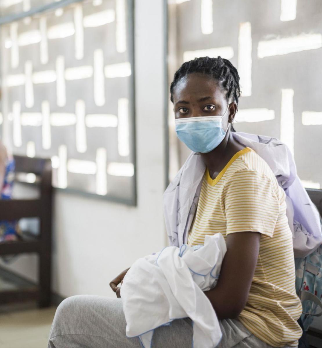 Une femme portant un masque de protection respiratoire et tenant son bébé dans les bras est assise dans une salle d'attente médicale avec d'autres femmes, le regard tourné vers l'objectif.