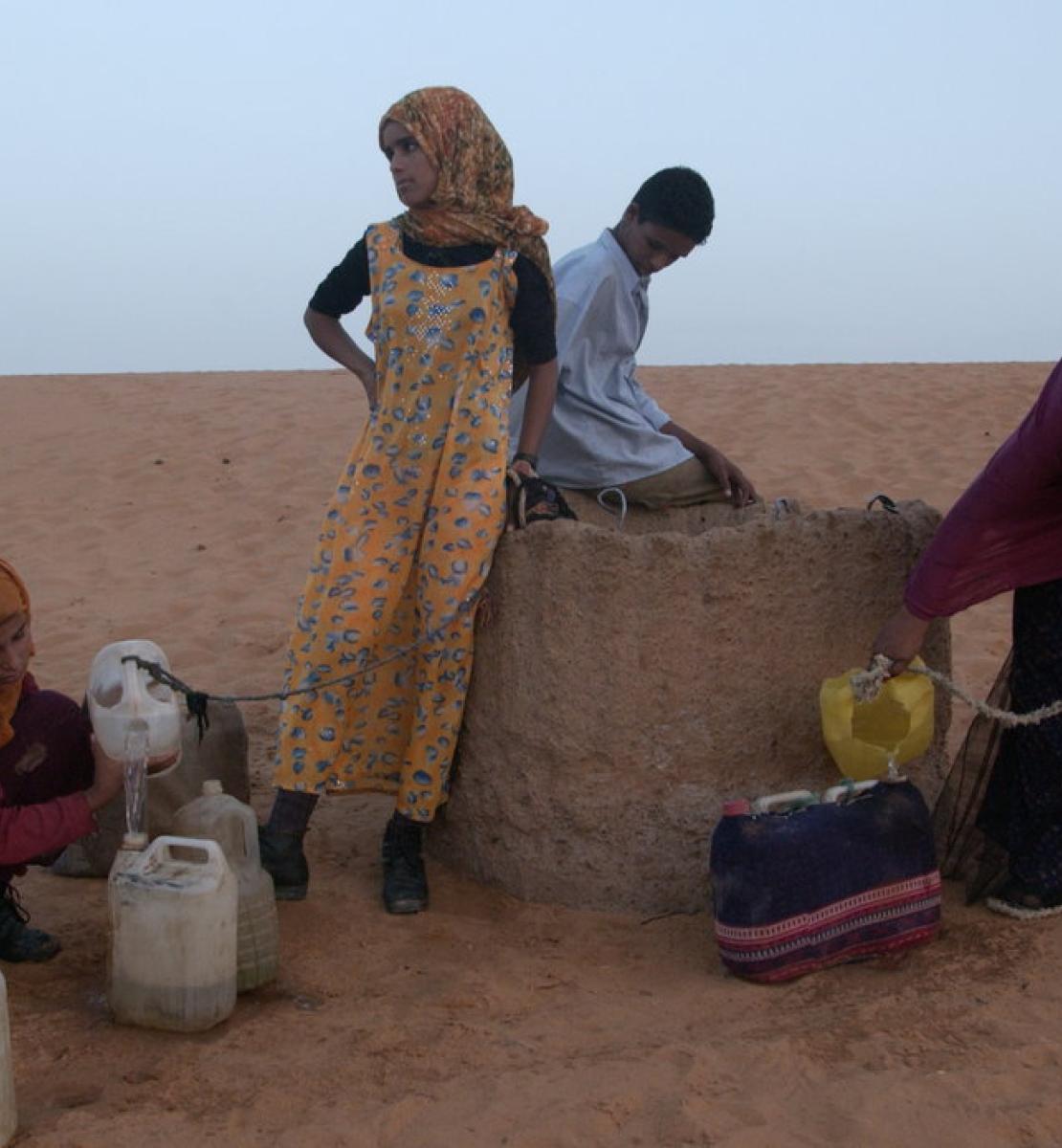 En un desierto de arena roja, cuatro jóvenes intentan llenar jarras de agua de un pozo.