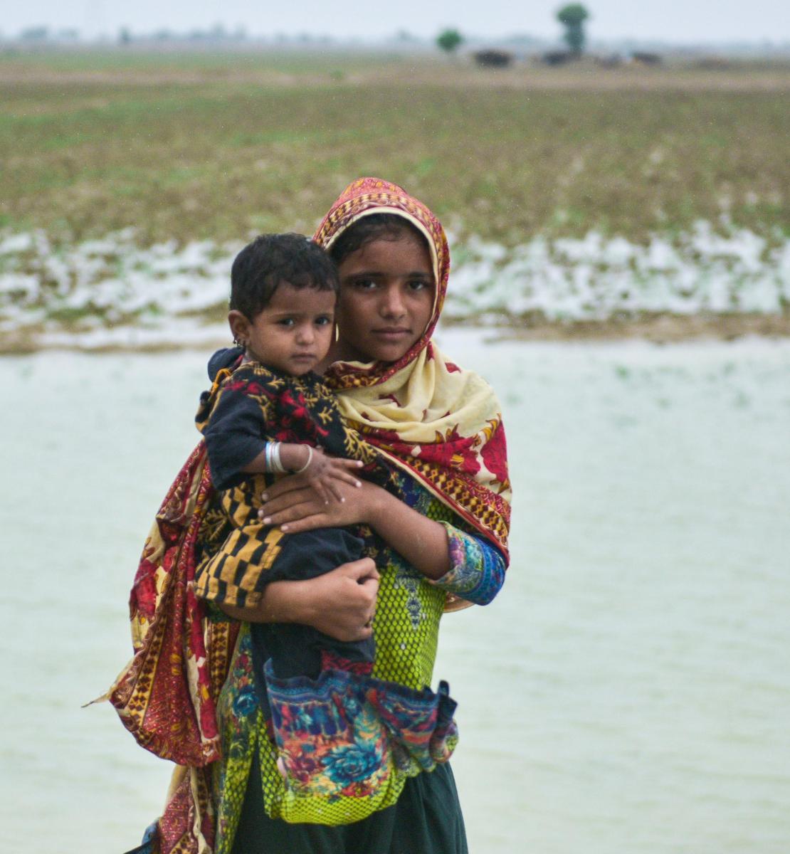 فتاة ترتدي وشاحًا تحمل طفلًا صغيرًا بين ذراعيها أمام بركة ماء.