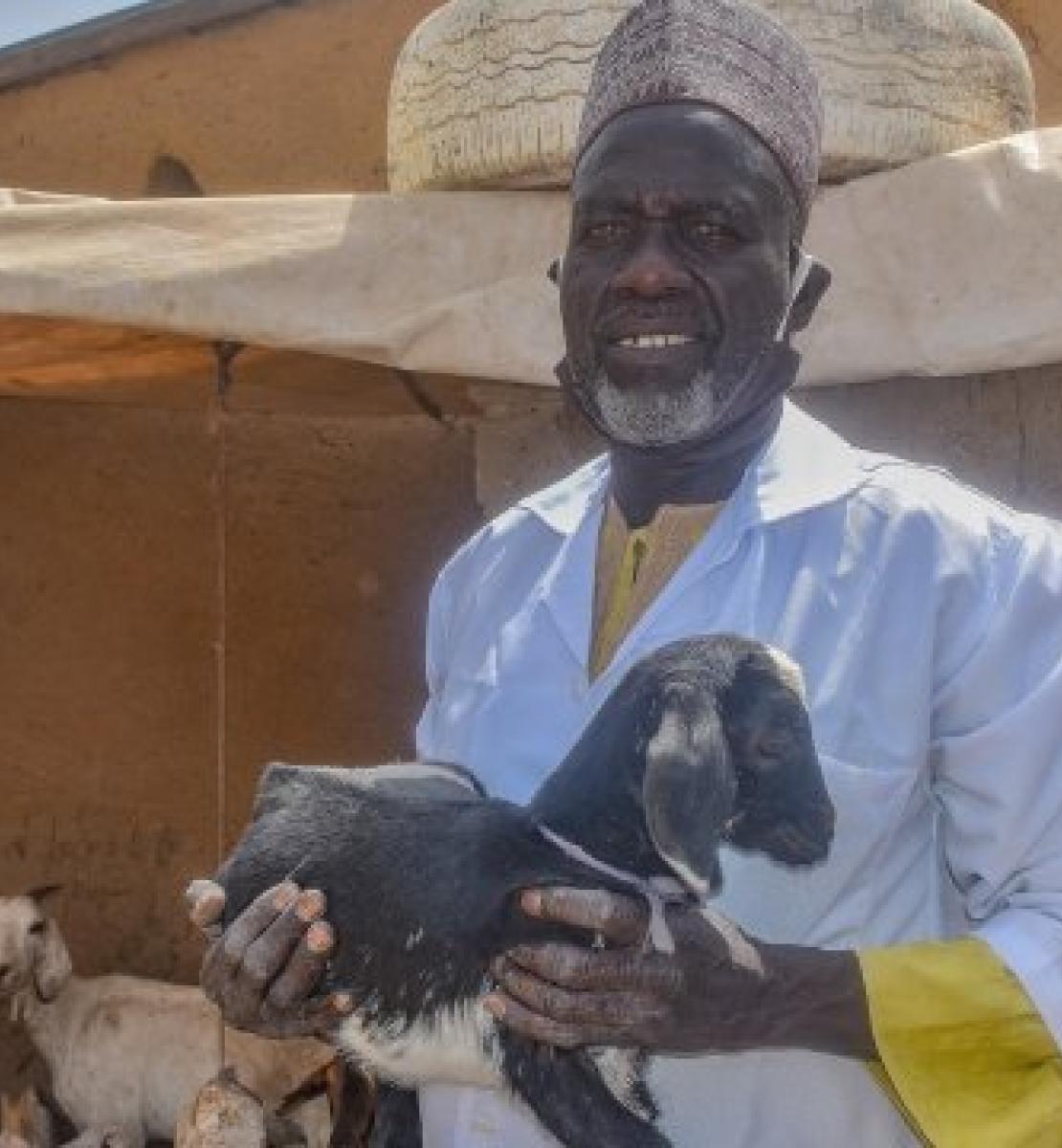 In Cameroon, a man wearing a sky-blue blouse and a head covering is photographed standing in front of a terracotta house, a baby goat in his arms, looking at the camera.