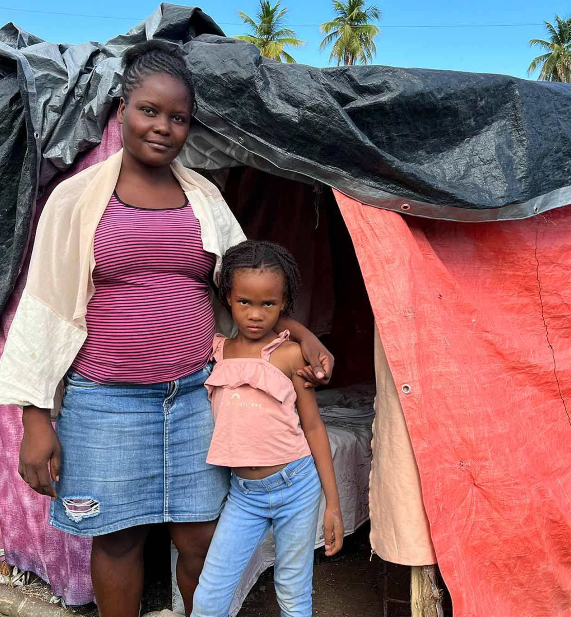 A young woman and her daughter stand in front of a temporary shelter covered with canvas and stare into the camera's lens on a sunny day in Haiti.