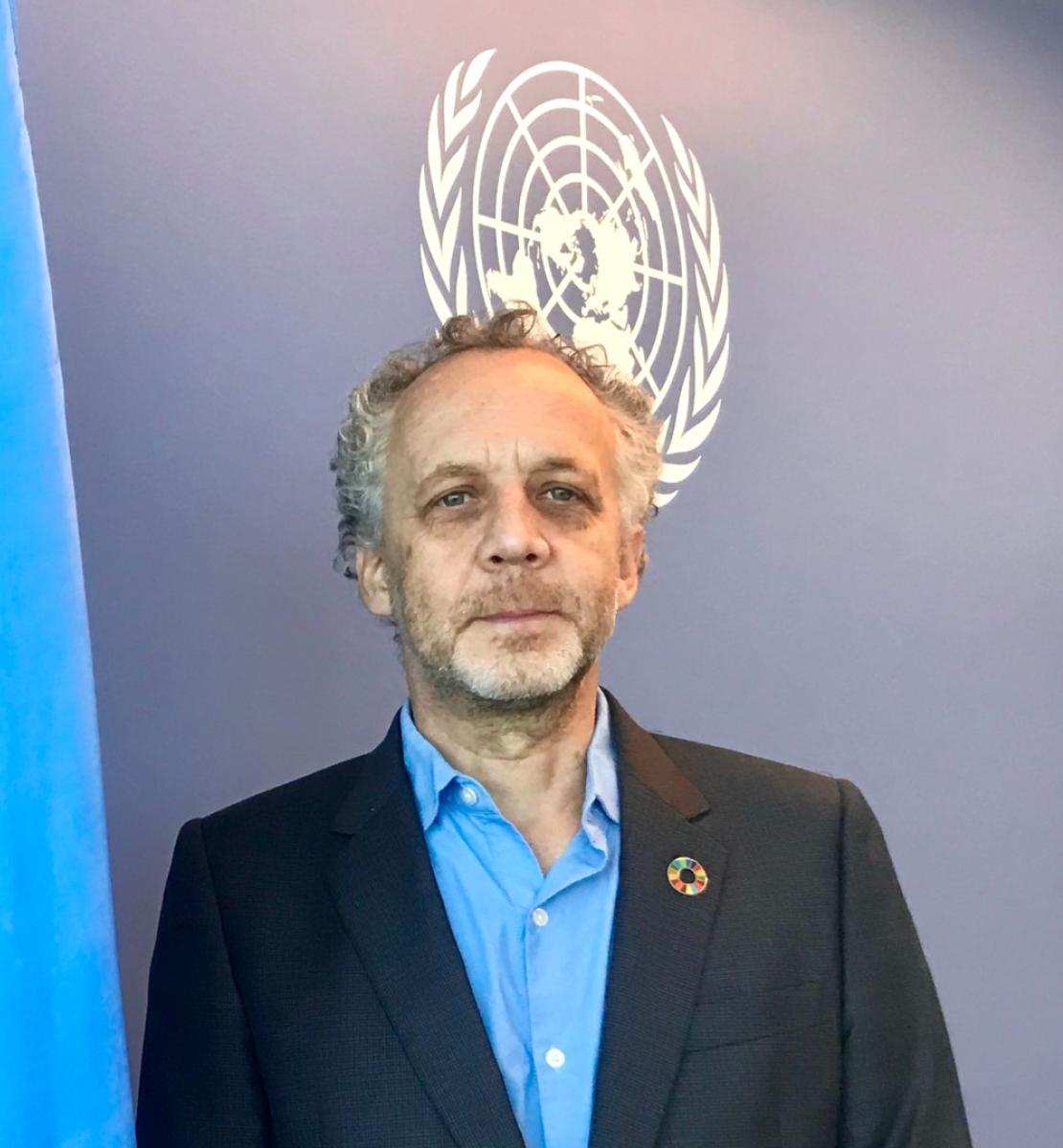 Joseph Scheuer, le nouveau Coordonnateur résident des Nations Unies au Cambodge, se tient devant le drapeau bleu de l'ONU, face caméra.
