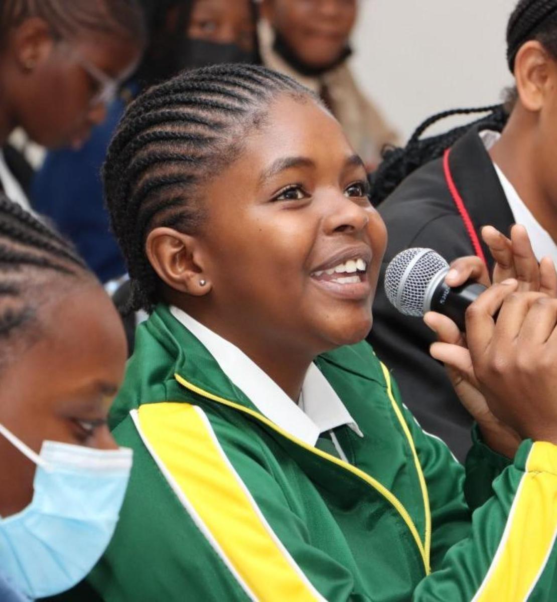 A young Namibian girl wearing a green tracksuit is sitting in a room with other girls and speaking at a microphone with a smile.