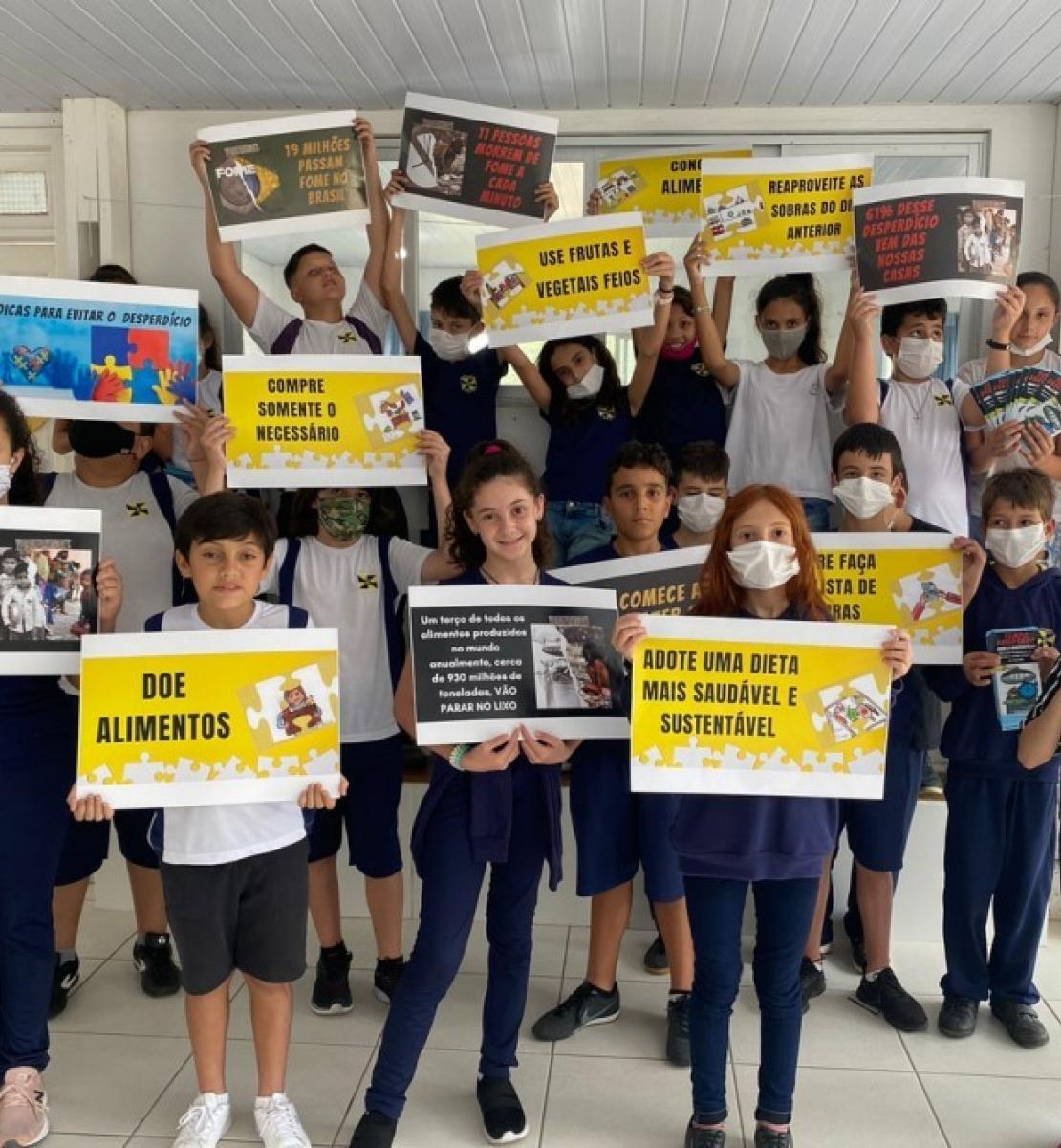 A group of students of elementary school age with their teacher. They are all wearing facemasks and standing side and proudly displaying their school work on waste management.