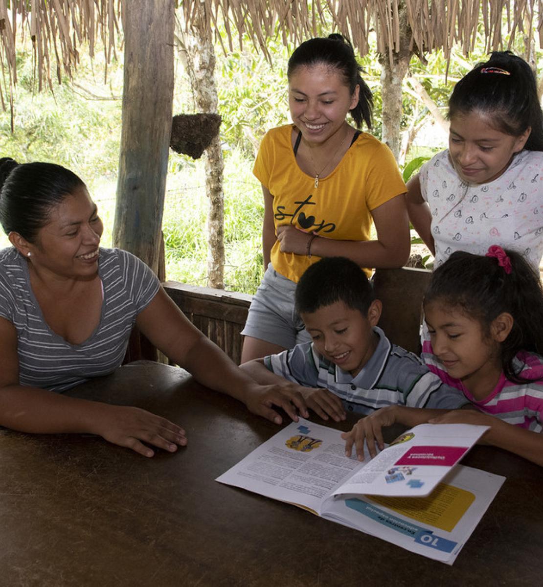 أربعة أطفال وامرأة يقرأون مواد تعليمية خلف طاولة خشبية.