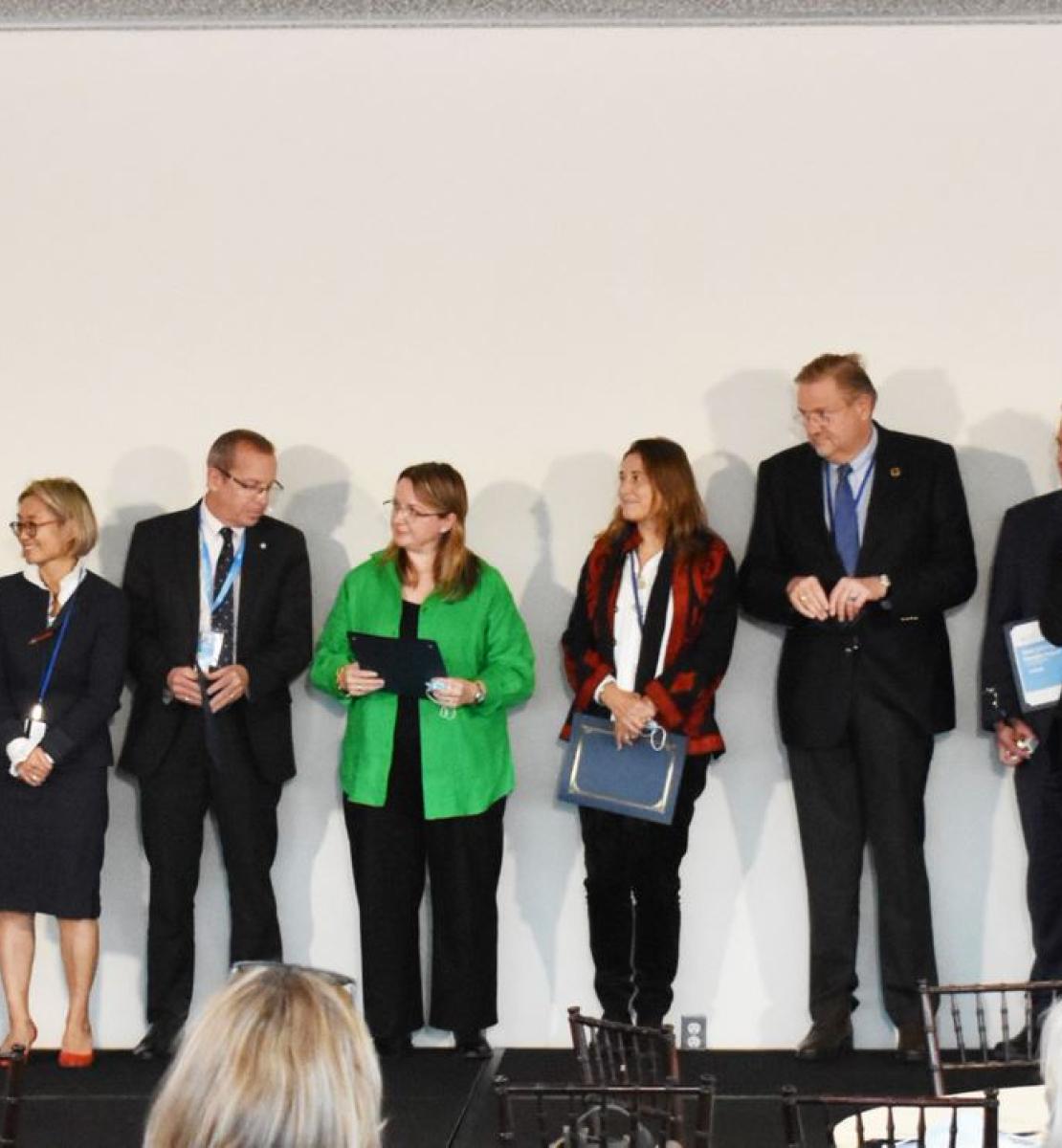 A group of sixteen people in business attire, standing next to each other, clapping and congratulating one another, on a stage during the UN Team Annual Results Reports Awards ceremony.