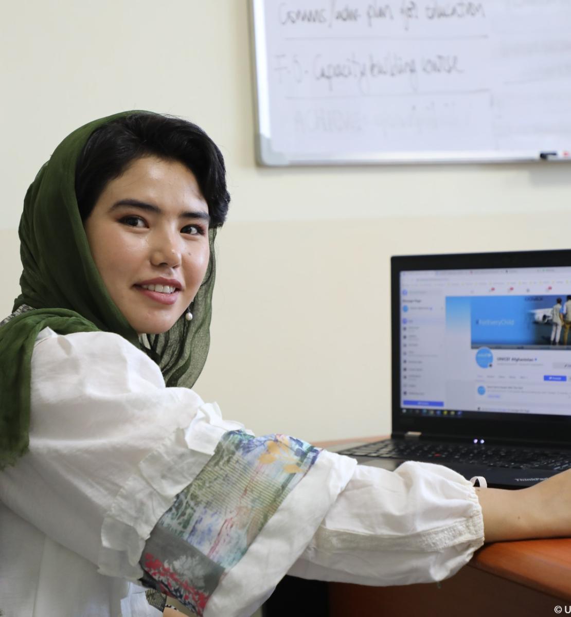 شابة أفغانية تبتسم للكاميرا وفي الخلفية جهاز كمبيوتر محمول على المكتب.