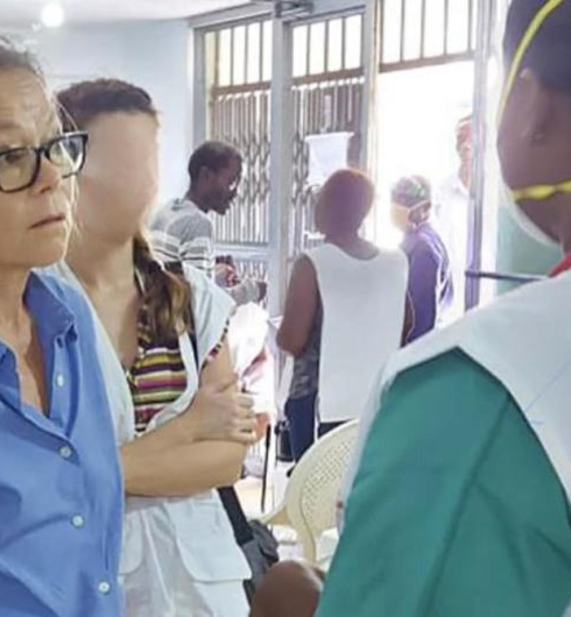 In a hospital in Haiti, a woman wearing a blue blouse and glasses talks with a caregiver photographed from behind, wearing a white gown and a respirator mask. The two women are surrounded by several other people. 
