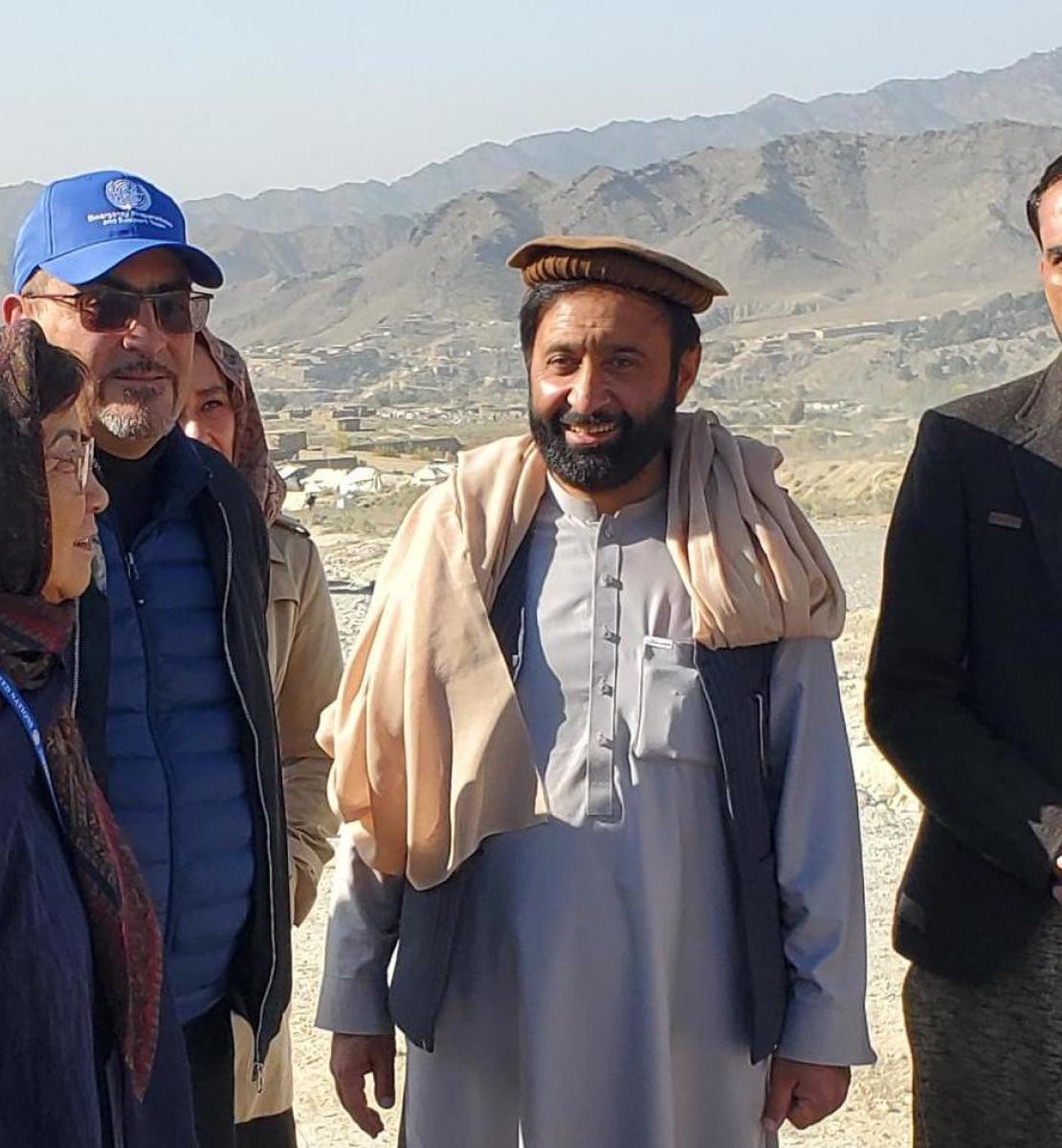A woman in a scarf stand beside a two men in caps and a man wearing a blazer, with tan mountains in the distance.