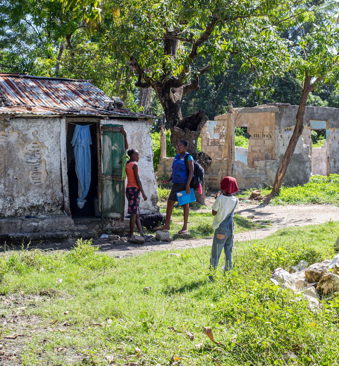 两名年轻妇女在一栋被树木包围的破旧房子的门口交谈。几个孩子正在附近玩耍。