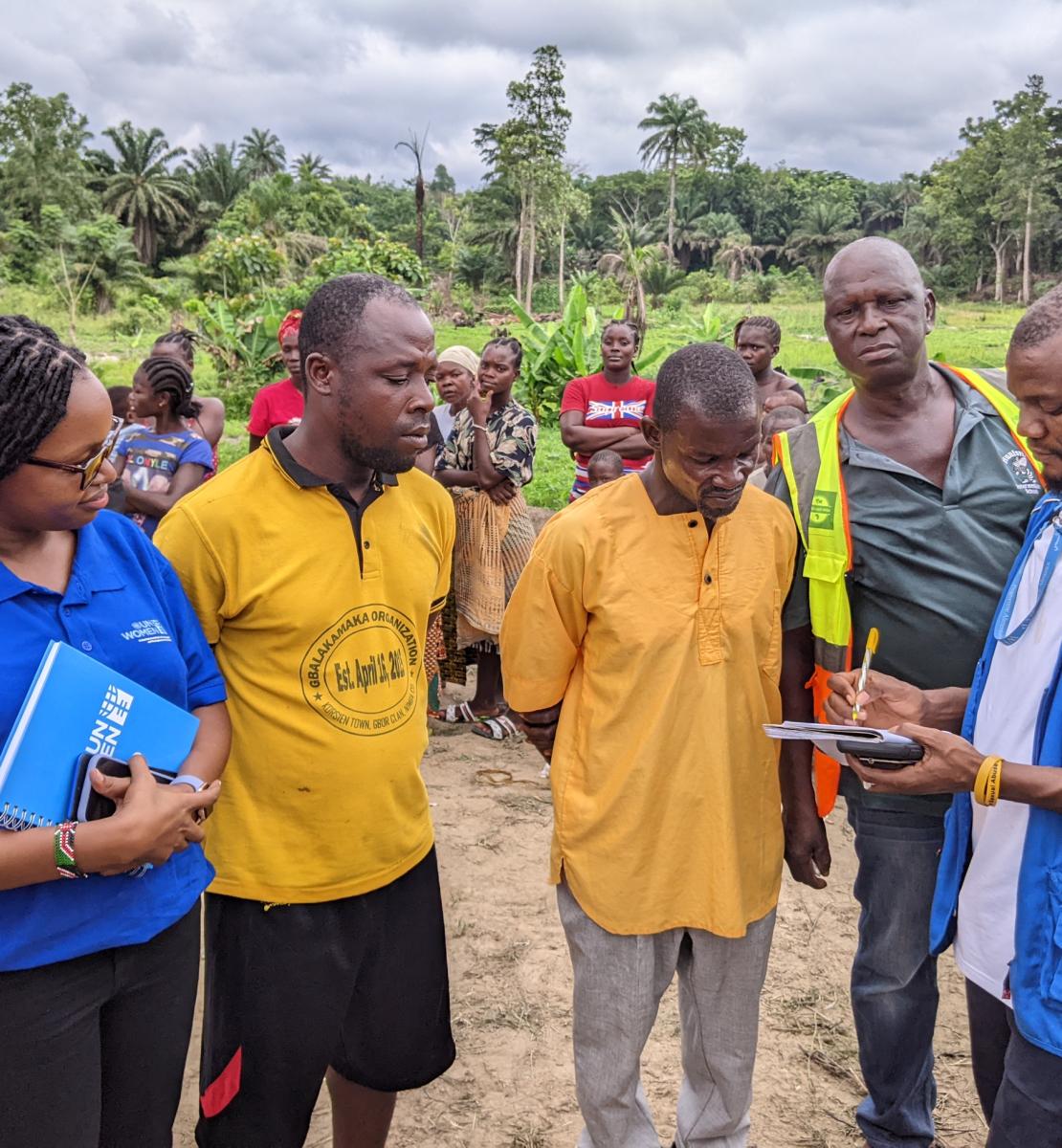 Au Libéria, à la lisière d'une forêt, deux hommes portant des t-shirts jaunes et une femme membre du personnel d'ONU Femmes regardent un troisième homme, membre du personnel du PAM, prendre des notes dans un cahier, tandis que plusieurs autres personnes se tiennent debout derrière eux.