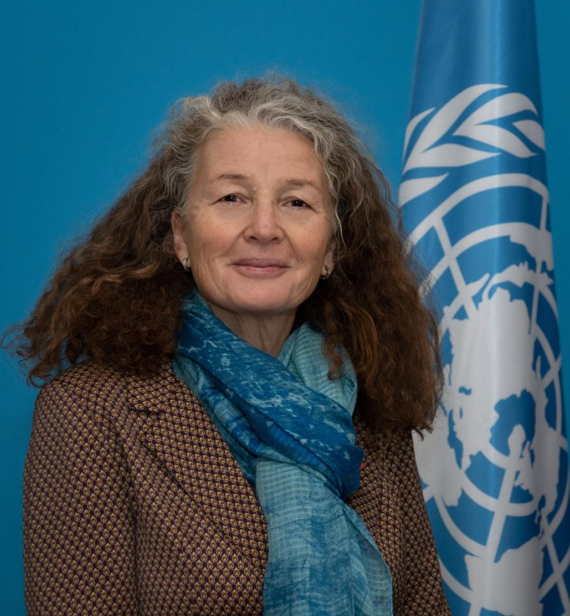 Une femme aux cheveux longs, bouclés et grisonnants et portant un blazer marron et une écharpe bleue se tient près d'un drapeau de l'ONU et regarde la caméra en souriant.