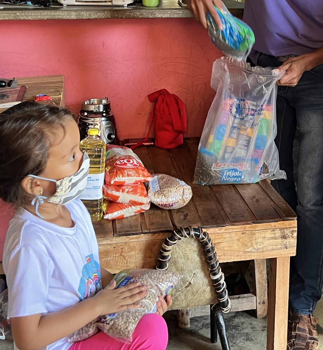 A small girl with a facemask holds a bag of beans while looking at two adults, and in the background there is a table with food on it.
