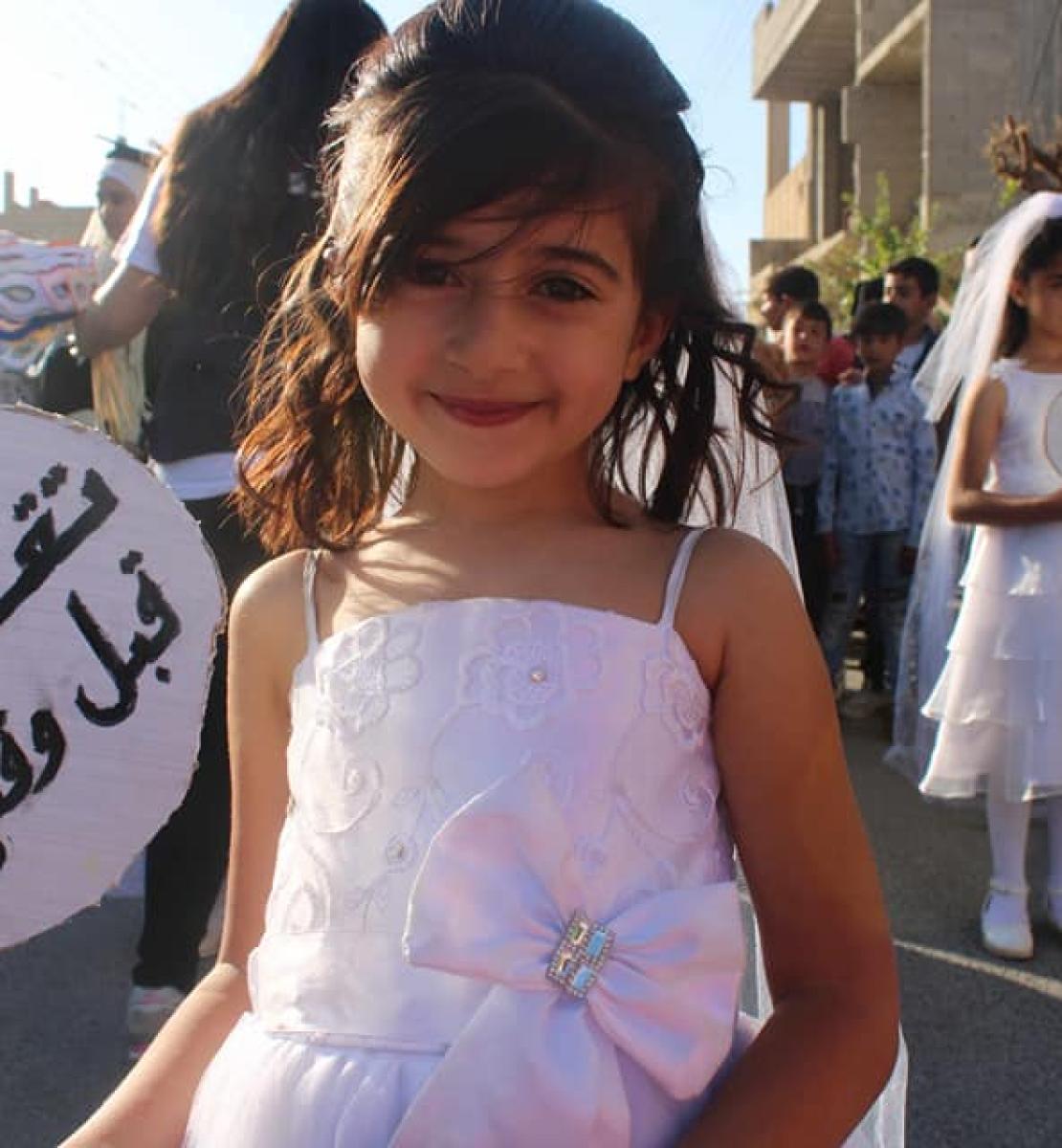 Young girl wearing a wedding dress and carrying a sign that says: don't marry her before her time.