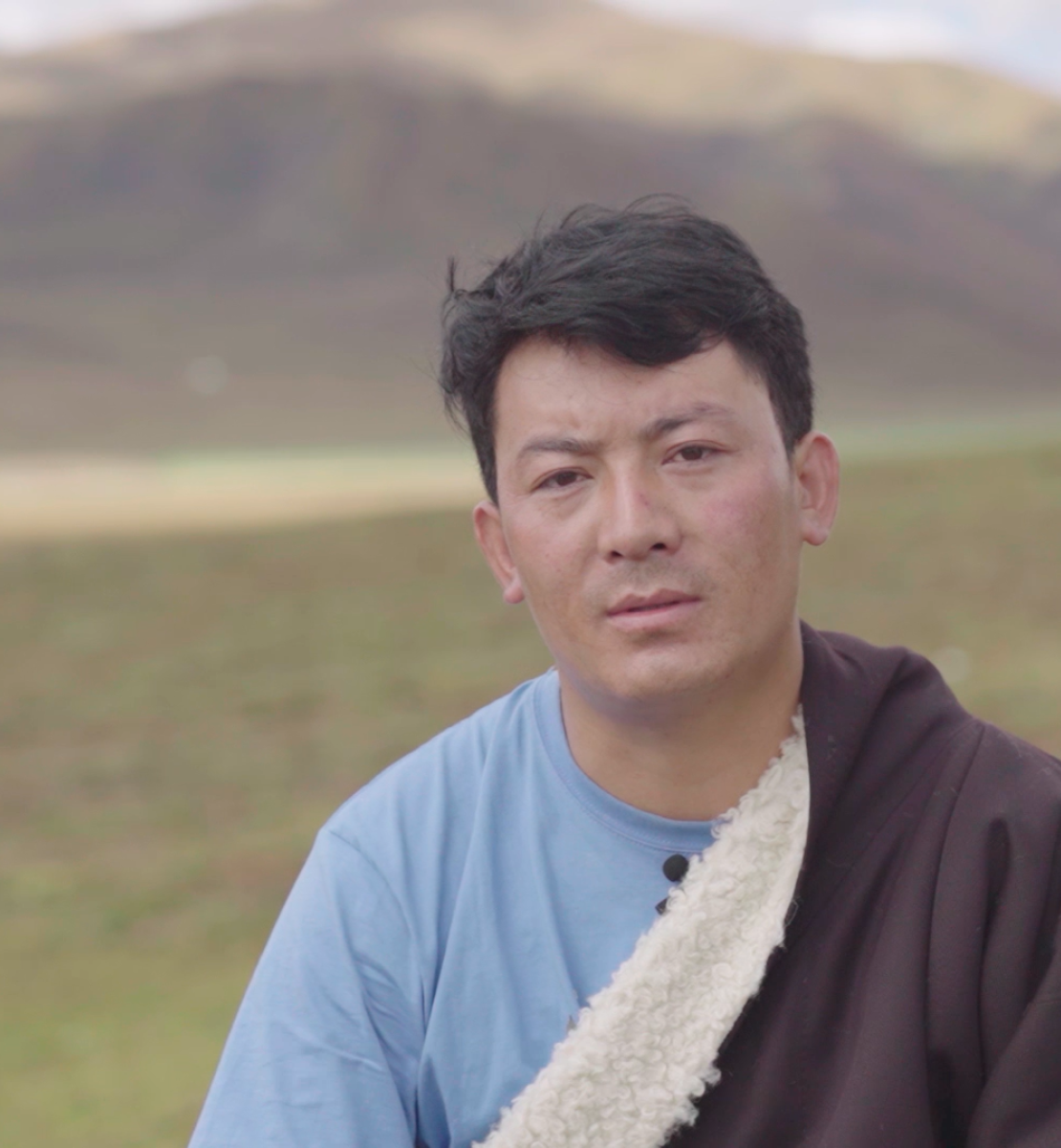 A Tibetan man sitting on grassland with mountains in the back. 