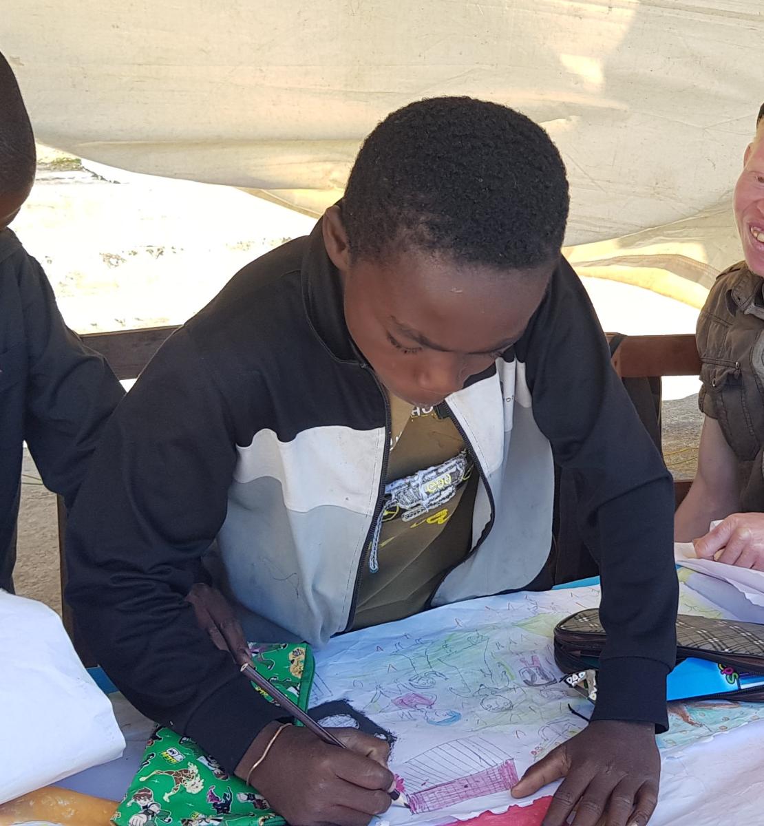 在马达加斯加，两个小男孩在联合国儿童基金会搭建的大帐篷下的桌子前学习，而第三个男孩，一个白化病患者，则带着灿烂的笑容看着。