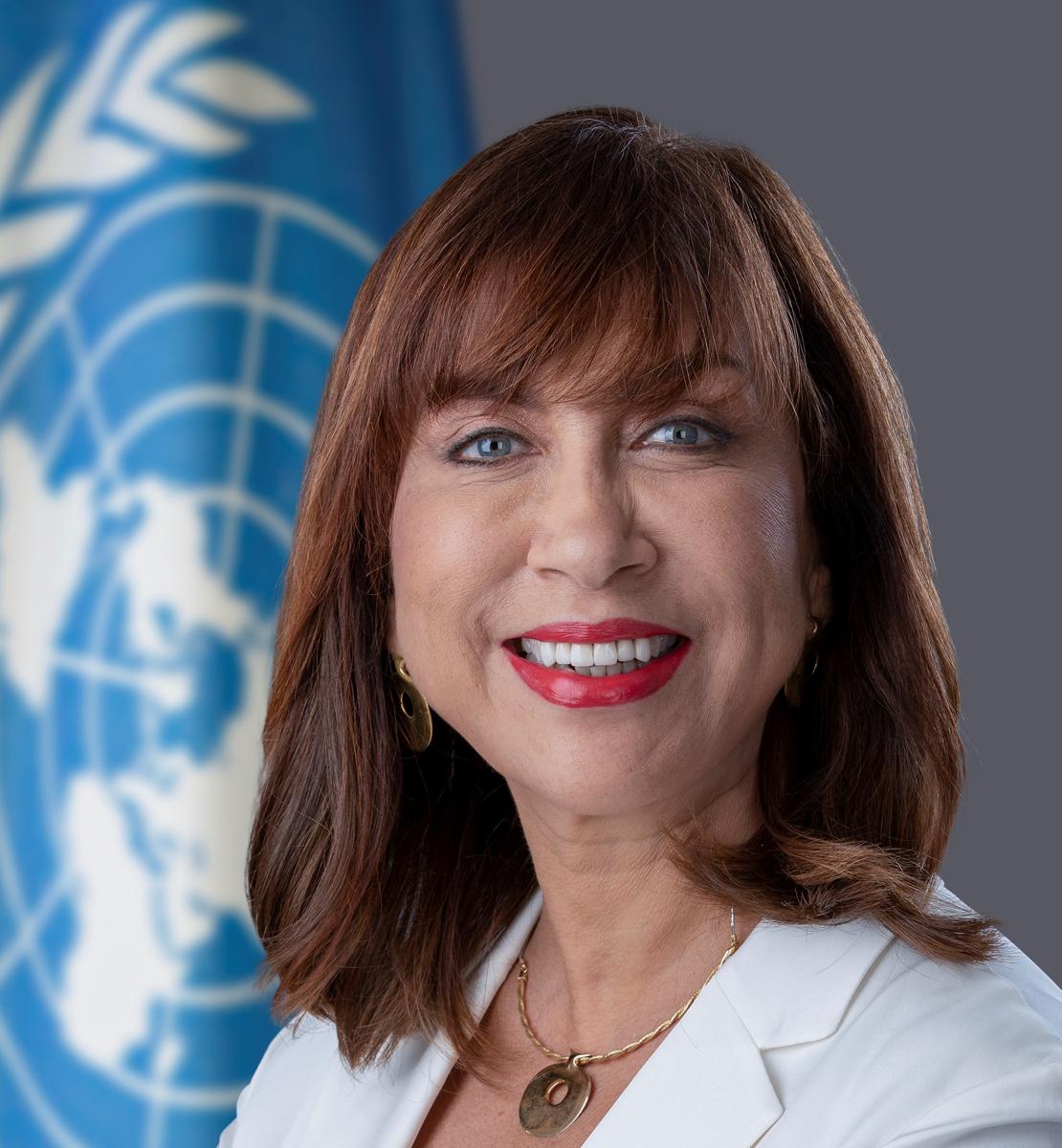 Women in white blazer smiles in front of UN flag 