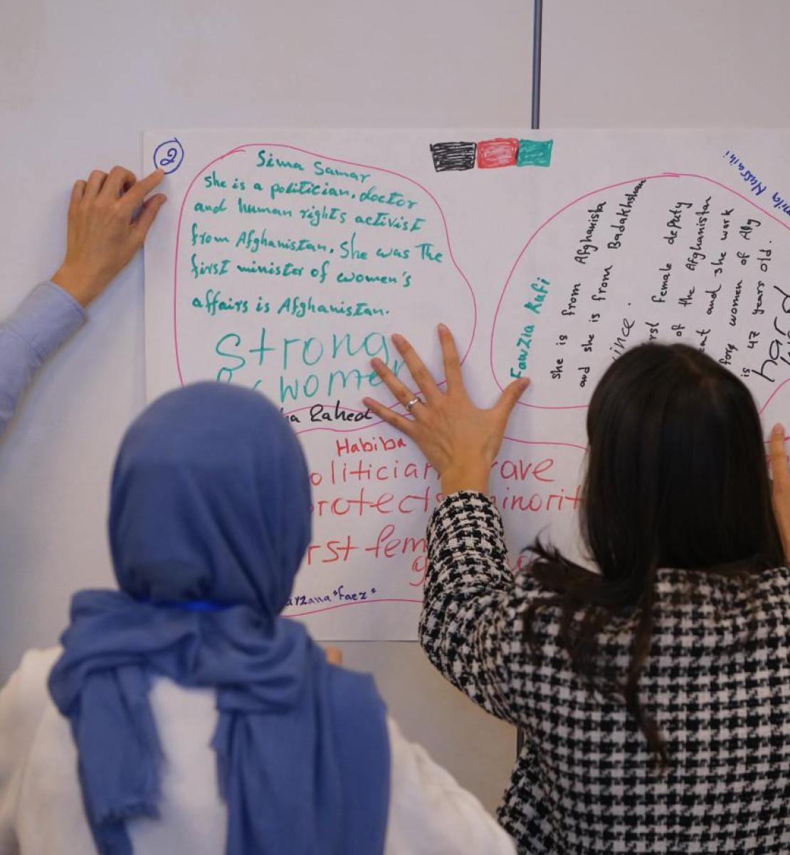 Three women work on a whiteboard and point to scribbled notes, with their backs facing the camera.