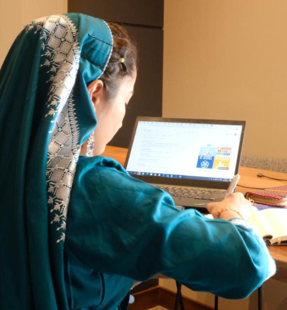 A female in a green satin-like dress with matching headscarf faces away from the camera and works on a laptop.