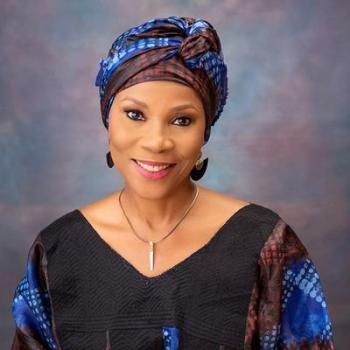A woman wearing a blue-patterned headwrap and a matching dress smiles and faces the camera. 
