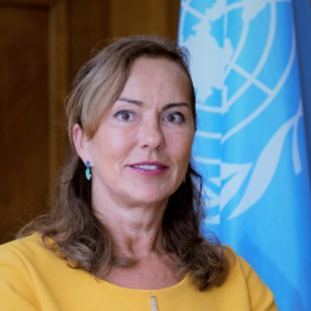 A woman in a yellow dress smiling in front of the United Nations Flag. 