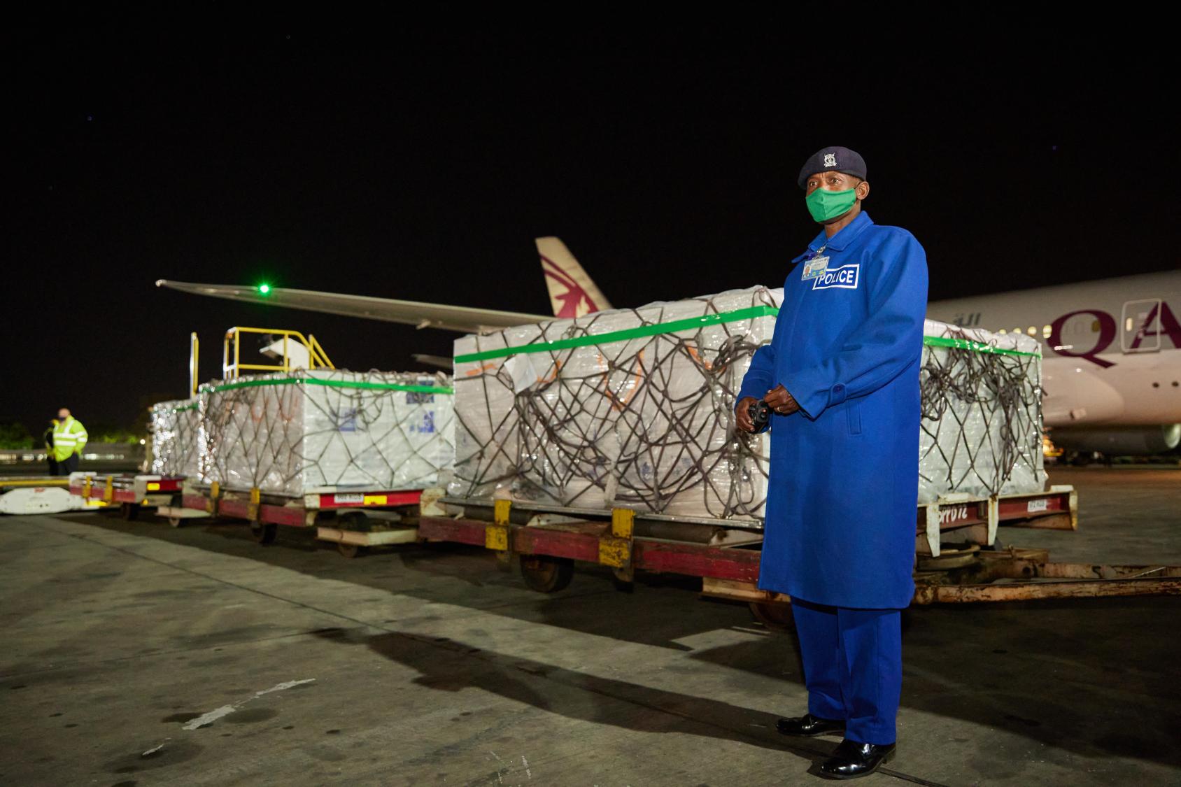 A man dressed in a blue police uniform stands next to a large delivery of vaccines with an airplane in the background.