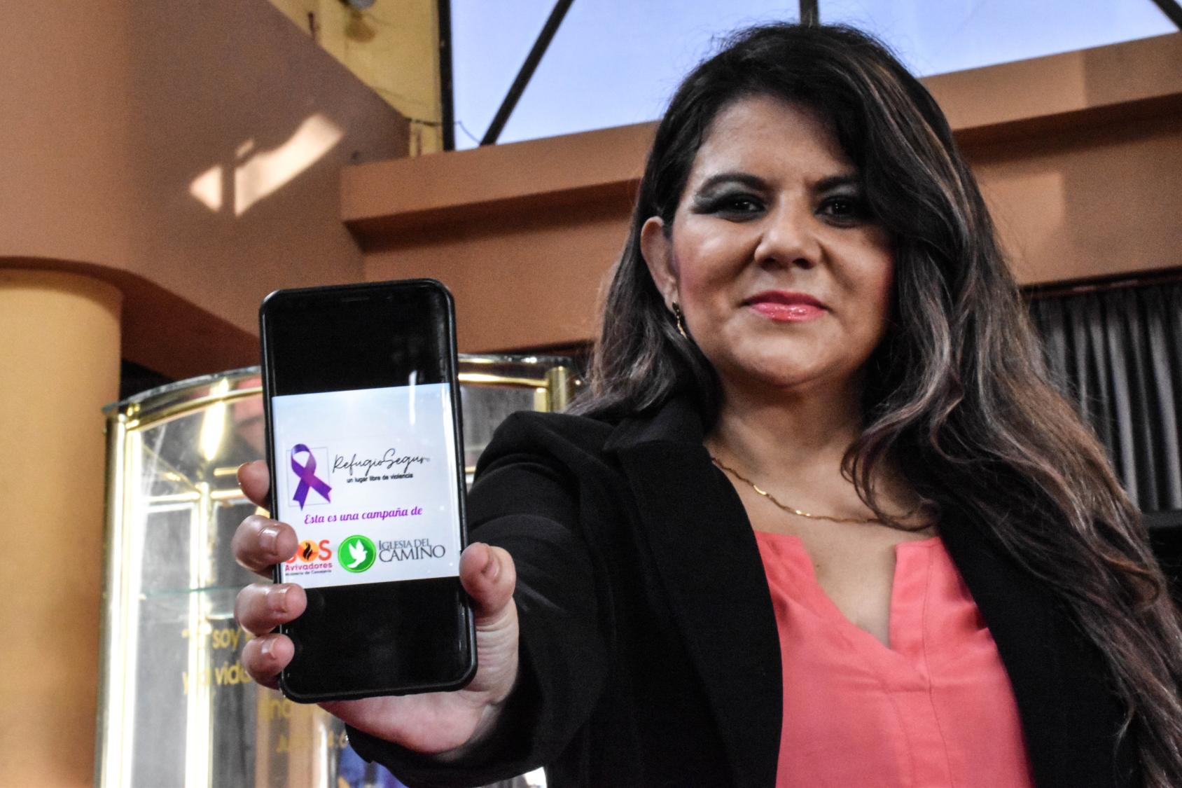 A woman facing the camera holds up a phone displaying a purple ribbon and words "Refugio Seguro." 
