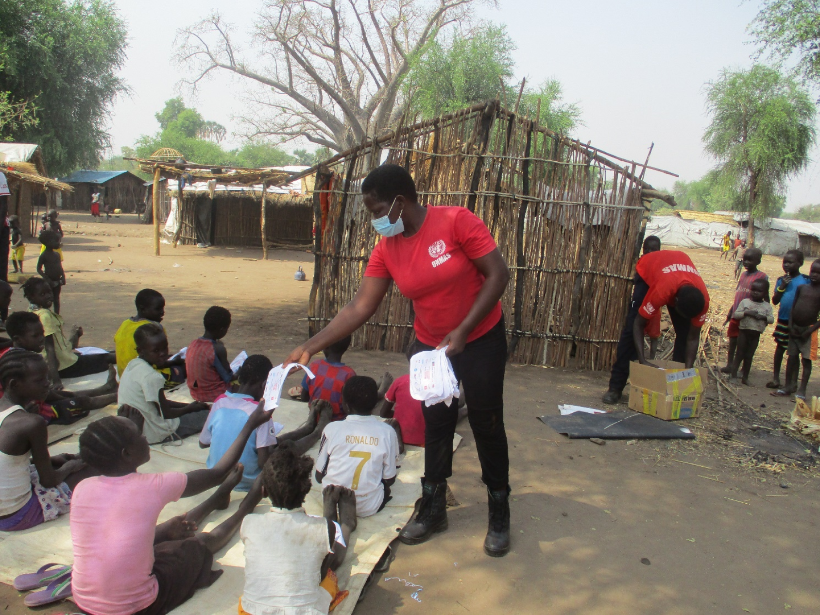 Женщина в красной рубашке и светло-голубой маске раздает информационные брошюры группе детей, сидящих на земле. 