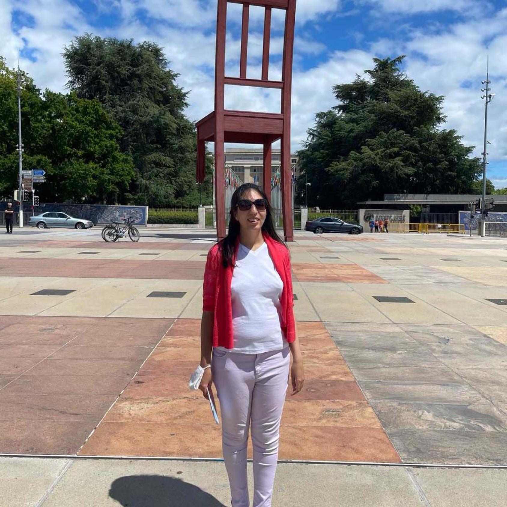 Samaneh wearing a red cardigan and a white shirt stands in front of a large statue of a chair. 
