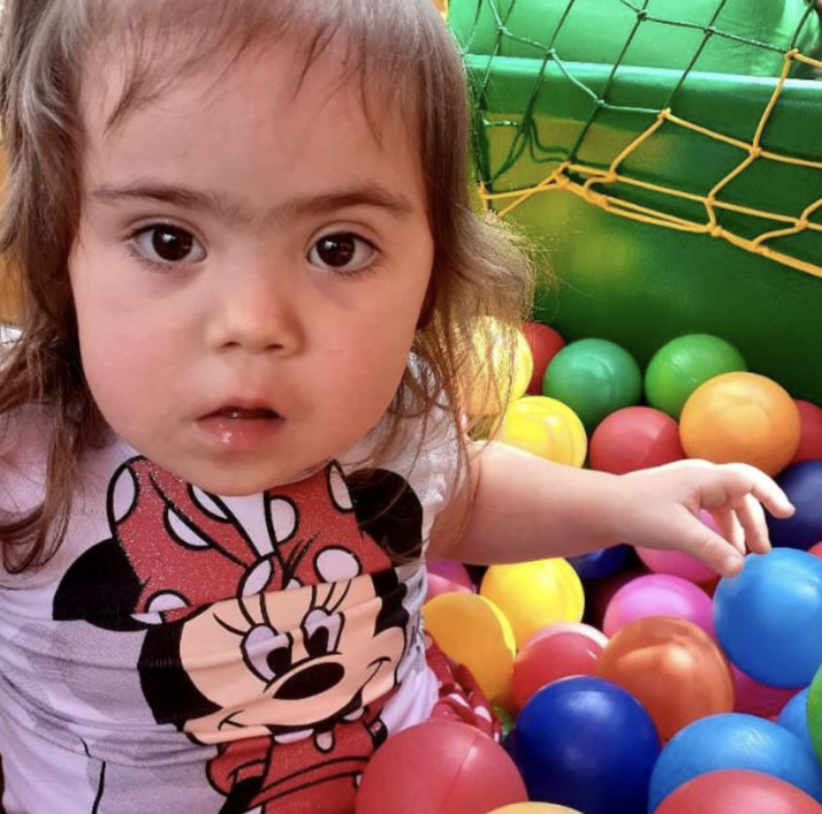 A little girl in a pink minnie mouse shirt sits in a ball pit with multi-colored balls. 