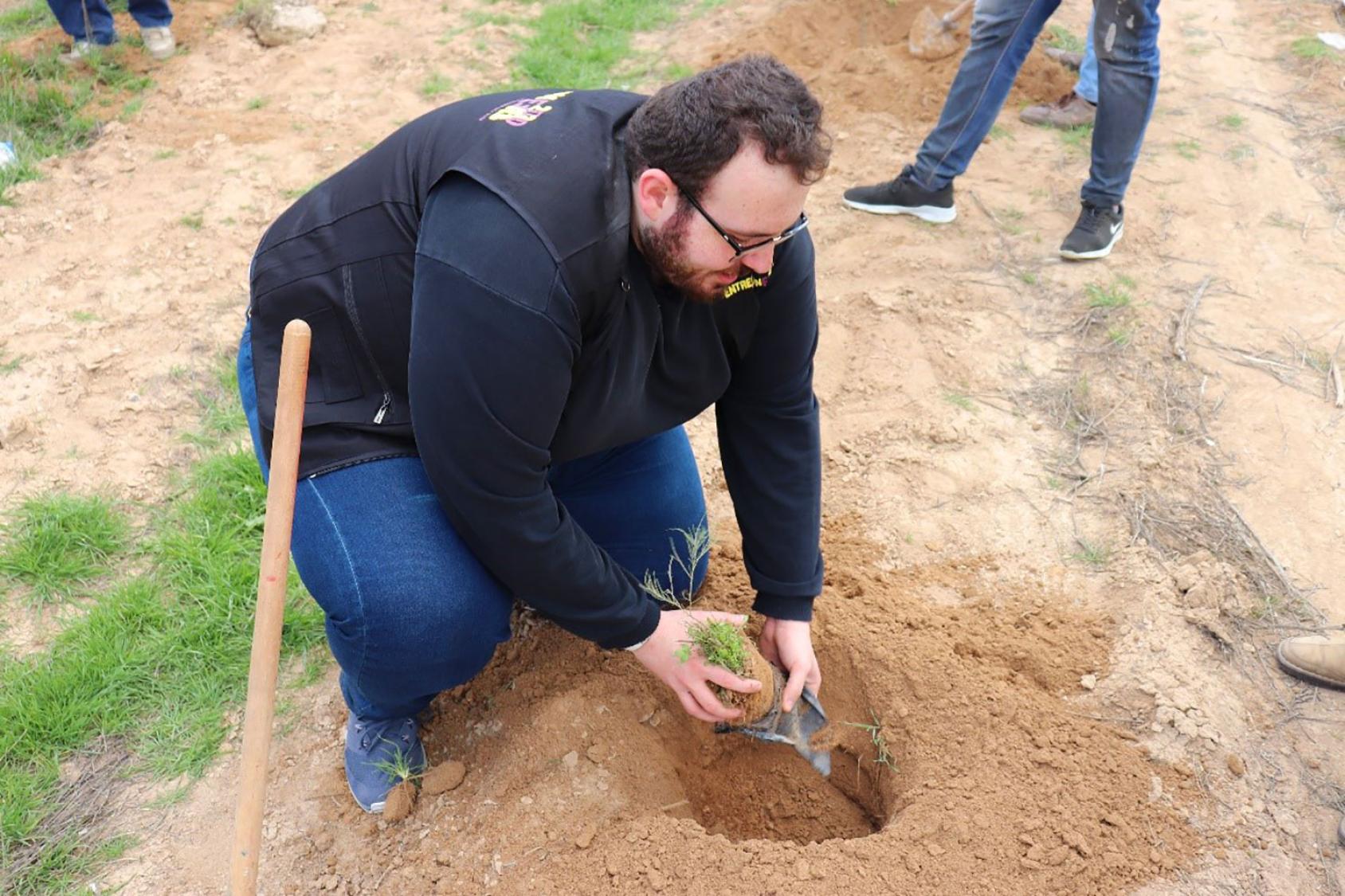 A man in a black shirt and black vest is kneeling near a hole where he is placing a tree.