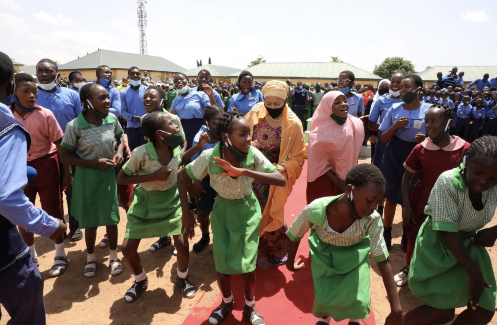 Several people stand together while children dressed in their green school uniform dance in the center of the group. 