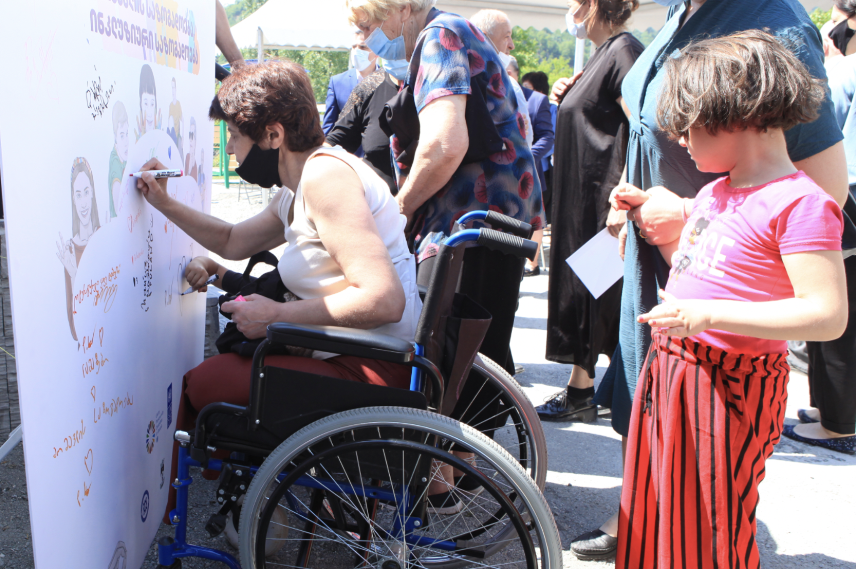 一位坐在轮椅上的妇女在海报板上签名，几个人站在附近。