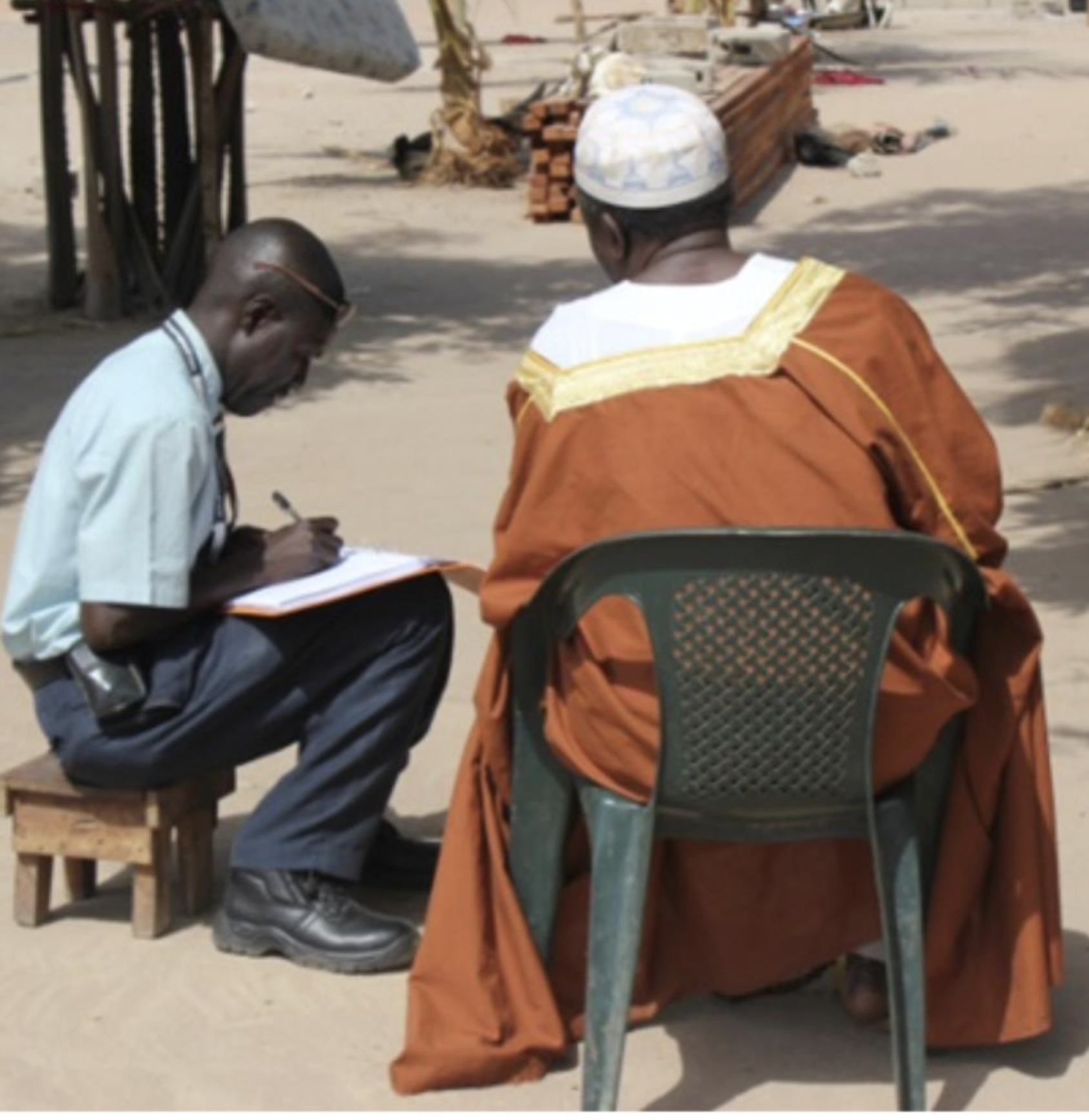 Un homme est assis sur un petit tabouret tandis qu'il prend des notes dans un cahier. Un deuxième homme est assis à côté de lui sur une chaise en plastique et répond à ses questions.