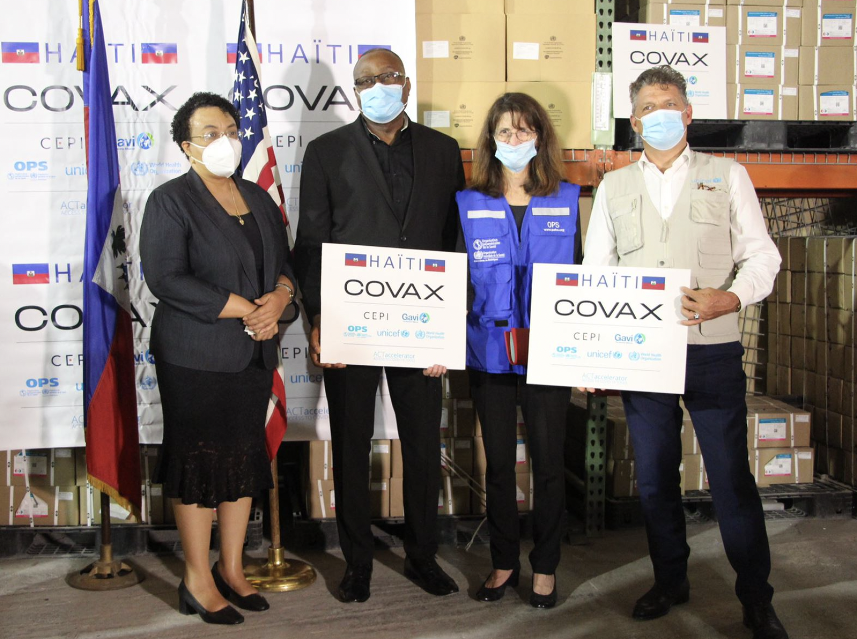 Four people hold COVAX signs in front of the flags of Haiti and United States.