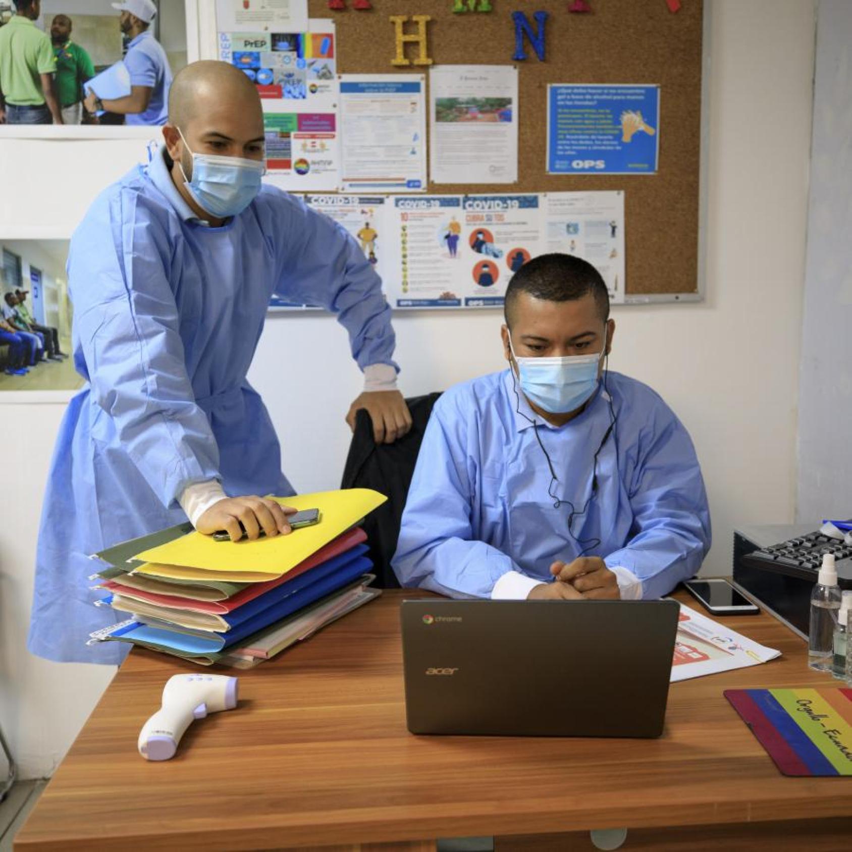 Two men in medical garb look at a computer on a desk. 