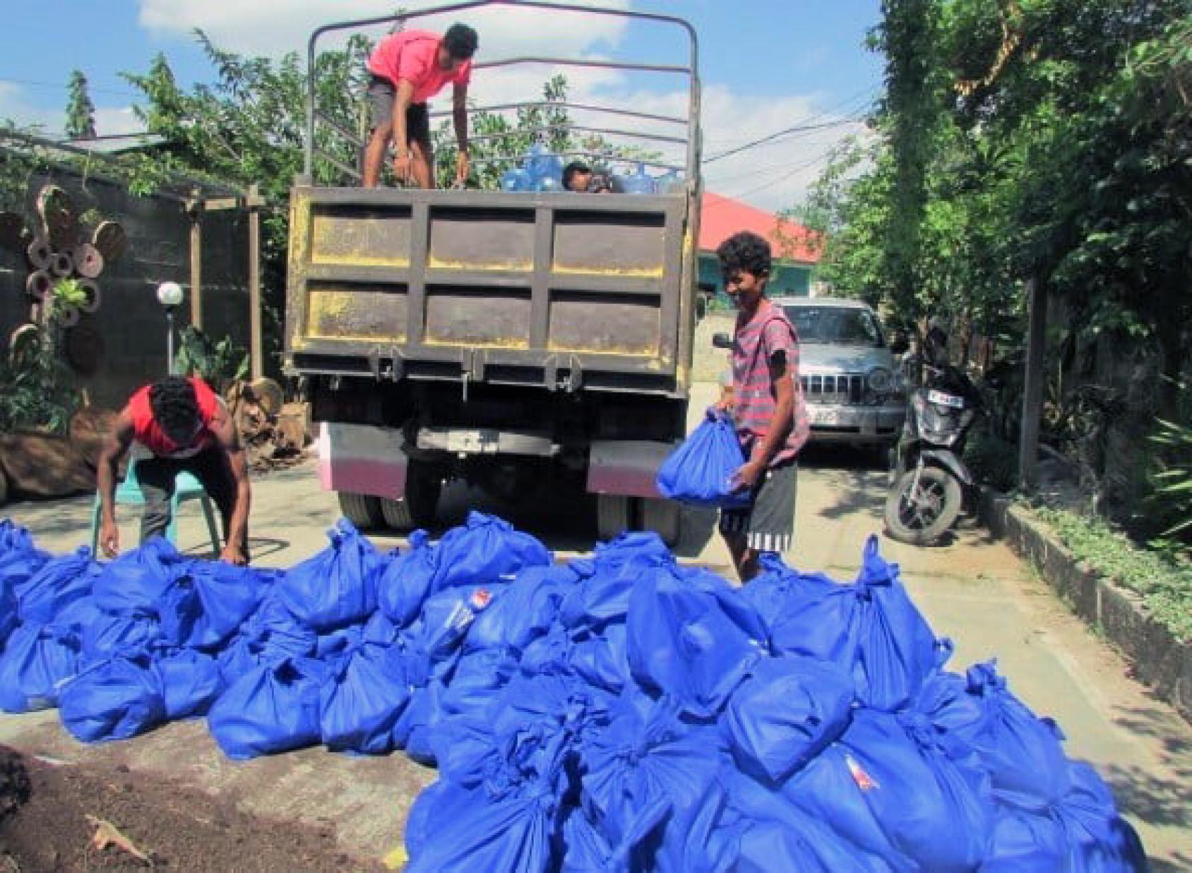 Dans une allée bordée d'arbres, trois jeunes hommes chargent un camion de gros sacs en plastique bleus contenant des produits de première nécessité destinés à être livrés aux personnes touchées par les inondations.