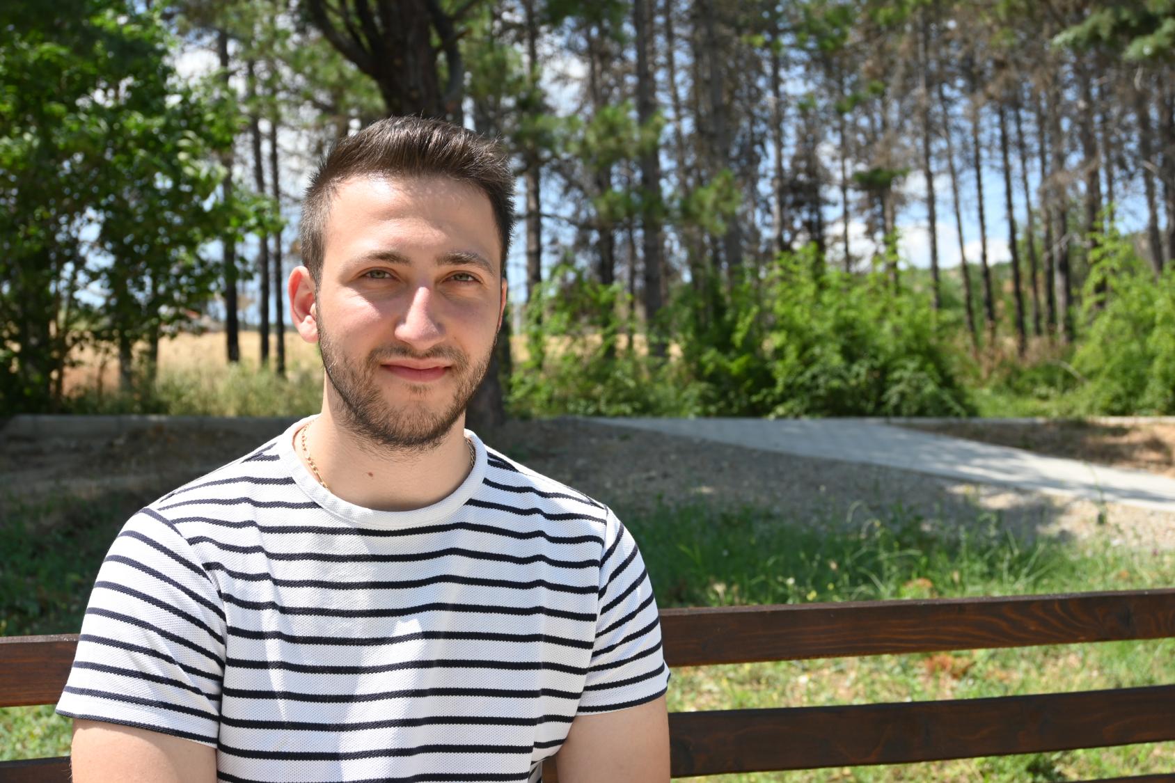 ألكسندر ميلوشفيك، المسؤول عن شبكة تثقيف الشباب على يد الأقران في مقدونيا الشمالية يظهر فيي يسار الصورة جالسًا على مقعد في حديقة.