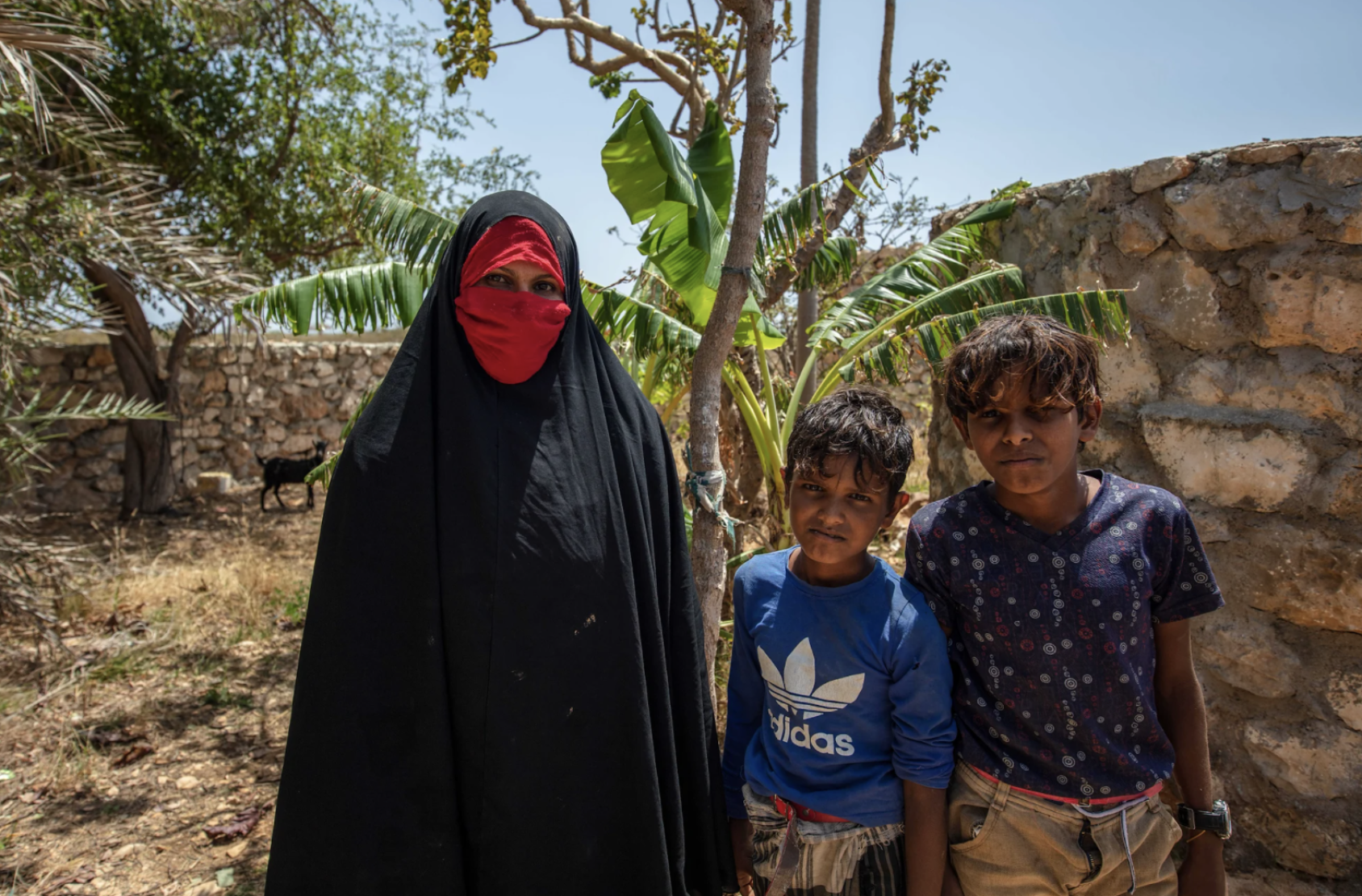 A woman in a black and red hijab stands next to two young boys in blue shirts. 