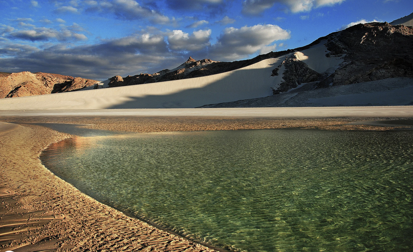 Image d'un étang au pied d'une colline de sable derrière laquelle s'élèvent des montagnes.