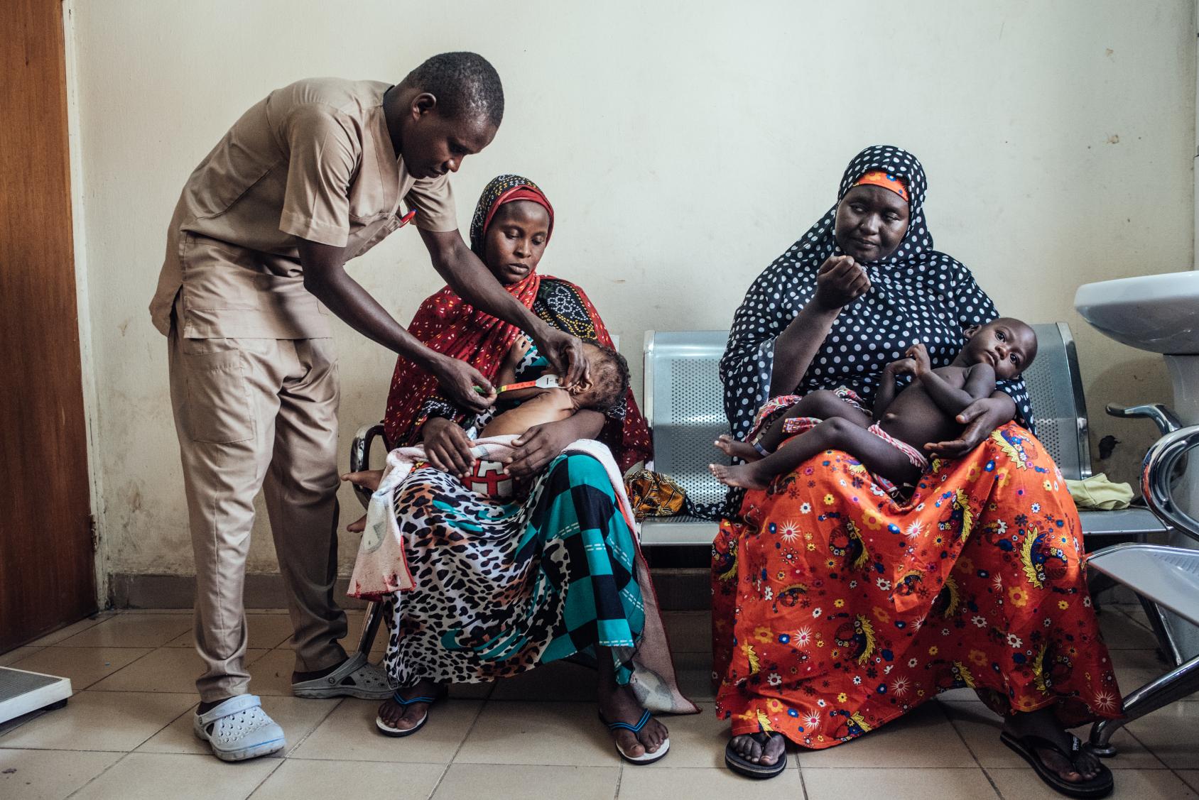 A healthcare professional attends to a malnourished child being held by their mother, while another woman sits next to them holding her child in a waiting room. 