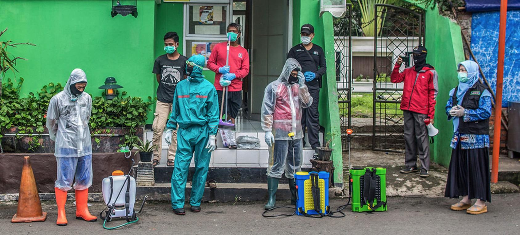 Plusieurs hommes portant un masque de protection respiratoire se tiennent devant la façade verte d'une maison. Les hommes photographiés au premier plan portent, chacun, un équipement de protection individuelle, leur matériel de désinfection posé au sol, près d'eux.