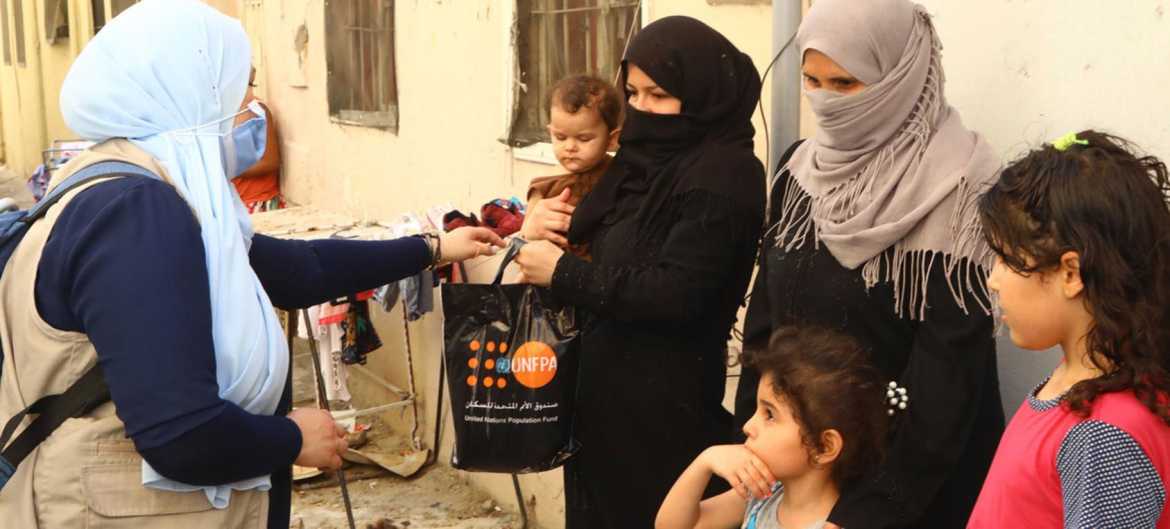 Un membre féminin du personnel de l'ONU portant un masque de protection remet un kit dignité du FNUAP dans un sac en plastique noir à une habitante de Beyrouth vêtue d'une burka et tenant un jeune enfant dans les bras. Une autre habitante se tient avec deux fillettes près de la femme en burka.