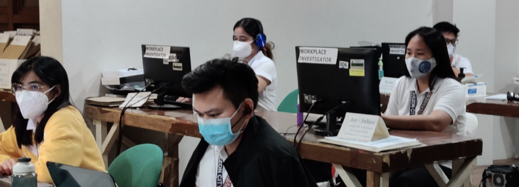 People in face masks sit inside a room with a computer in front of each person. 