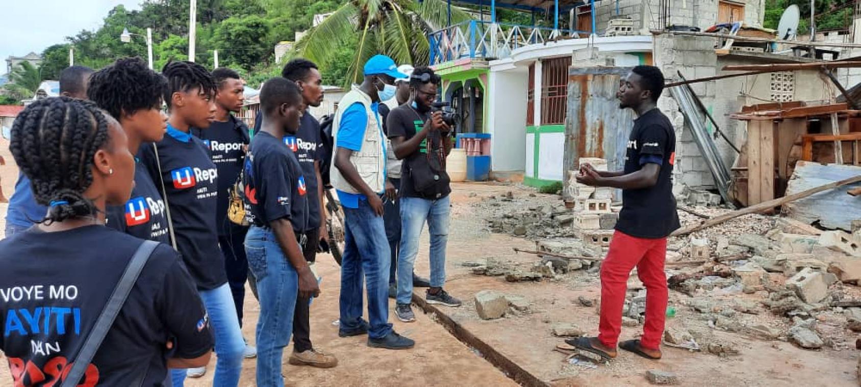 Un jeune homme en t-shirt noir et pantalon rouge s'adresse à plusieurs jeunes volontaires U-Reporters et travailleurs humanitaires au milieu des décombres causés par le tremblement de terre qui a frappé Haïti en août 2021. Un des jeunes présents dans le groupe le filme en train de parler à l'aide de sa caméra.