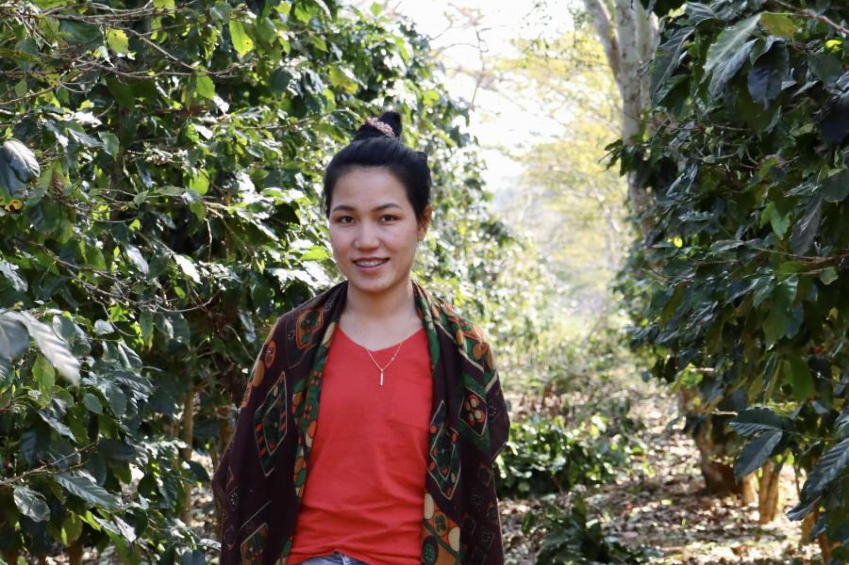 A woman, in a red shirt, smiles at the camera in a coffee field. 